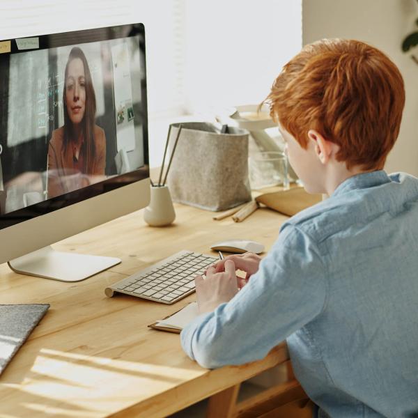 A redheaded student in a blue shirt sits at a comfortable desk interacting with a teacher