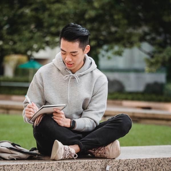 A high school age boy with black hair and a sweatshirt sits on a wall with a notebook