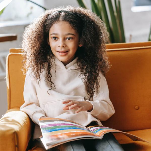 A child with long very curly brown hair sits on a sofa with a workbook