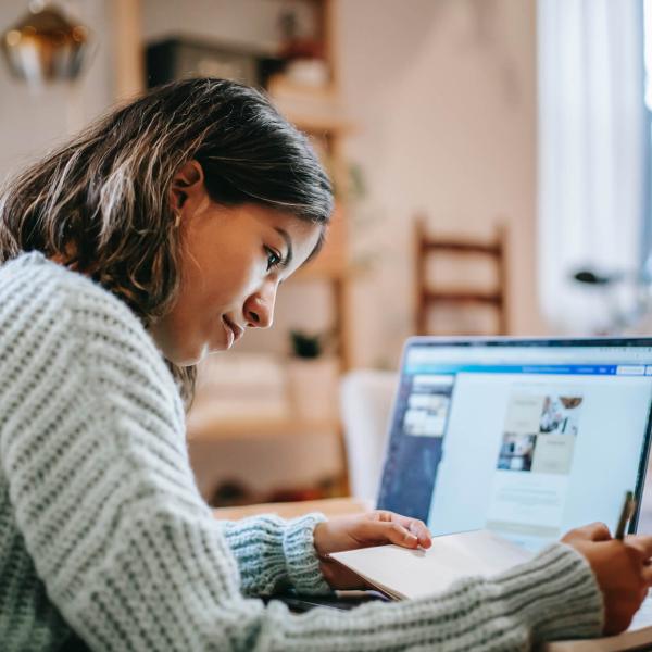 A high school age woman with black hair takes note. Her laptop is open in the background.