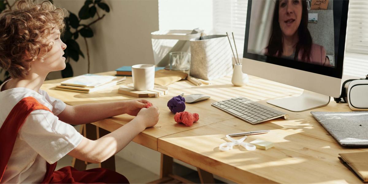 Child with curly red hair and red overalls works with moldable clay while talking with teacher on computer monitor